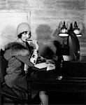 1920s SMILING WOMAN WITH PEN TO LIPS WRITING A LETTER AT DESK WITH LAMP IN HOTEL LOBBY WEARING CLOCHE HAT AND FUR COLLAR COAT