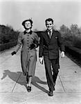 ANNÉES 1940 SOURIANTE JEUNE ADOLESCENTE COUPLE MARCHANT SUR LE TROTTOIR SOURIANT HABILLÉ ENDIMANCHÉS