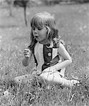 1960s GIRL SITTING IN FIELD BLOWING ON DEAD DANDELION