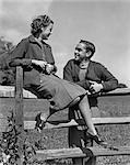 1930s 1940s YOUNG SMILING TEENAGE COUPLE TALKING GIRL SITTING ON WOODEN SPLIT RAIL FENCE