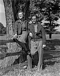 1940s TEEN COUPLE BOY GIRL HAPPY SMILING FULL LENGTH PORTRAIT BY WOODEN FENCE AND TREE