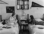 1930s 1940s TWO TEEN GIRLS LYING ON DORMITORY BEDS ROOM MATES LISTENING TO RADIO COLLEGE SCHOOL PENNANTS ON WALL