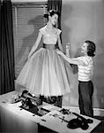 1950s TWO TEEN GIRLS WITH SEWING MACHINE ONE GIRL MODELING FANCY PROM DRESS OTHER GIRL ADJUSTING WAISTBAND