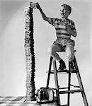 1950s SMILING BOY ON LADDER STACKING UP LARGE TOWERING PILE OF TOASTED BREAD