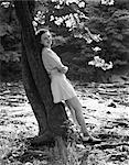 1940s SMILING YOUNG WOMAN WEARING SHORT SKIRT AND SADDLE SHOES LEANING AGAINST TREE BY STREAM