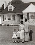 1950s FAMILY OF FIVE WITH BACKS TO CAMERA ON LAWN LOOKING AT FIELDSTONE HOUSE