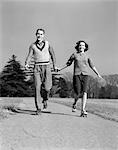 1940s SMILING TEENAGE COUPLE BOY GIRL HOLDING HANDS ROLLER SKATING ON SIDEWALK