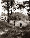 1930s REAR VIEW OF BOY & GIRL IN STRAW HATS & OVERALLS WALKING DOWN FARM ROAD WITH DOG BETWEEN THEM