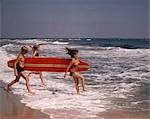 1970s ONE TEENAGE BOY TWO GIRLS IN BATHING SUITS LAUGHING RUNNING INTO OCEAN SURF CARRYING A RED SURFBOARD