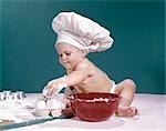 1960s BABY WEARING CHEFS HAT HOLDING EGG SURROUNDED BY COOKING EQUIPMENT