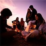 1970s TEENAGERS GATHERED AROUND BEACH CAMPFIRE PLAYING ACOUSTIC GUITARS