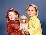 LITTLE BOY AND GIRL IN RAINCOATS AND HATS HOLDING COCKER SPANIEL PUPPY STUDIO