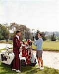 1960s SMILING COUPLE MAN WOMAN SELECTING GOLF CLUBS FROM GOLF BAG ON CART BY SAND TRAP