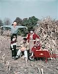 1950s FAMILY MAN FATHER WOMAN MOTHER BOY SON GIRL DAUGHTER IN AUTUMN CORN FIELD CARVING PUMPKIN SHOCKS SCARECROW RED WHEEL BARROW