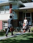 1950s WOMAN MOTHER GIRL DAUGHTER WAITING AT FRONT STEPS SUBURBAN HOUSE FOR MAN FATHER WITH BRIEFCASE COMING HOME