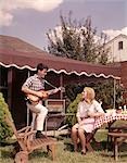 1960s TEENAGE BOY AND GIRL AT PICNIC TABLE LUNCH IN SUBURBAN BACKYARD BOY PLAYING ACOUSTIC GUITAR