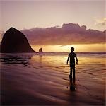 SILHOUETTED BOY WALKING ON BEACH AT SUNSET HAYSTACK ROCK NEAR CANNON BEACH OREGON