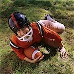 1970s FRECKLE FACED BOY FOOTBALL UNIFORM HELMET ORANGE TOP LYING IN GRASS HOLDING FOOTBALL LOOKING UP