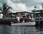 1950ER JAHRE FAMILIE BE-PICKNICK IN KLEINEN HÖLZERNEN MOTOR BOOT SCHLAUCHBOOT MOM DAD JUNGE MÄDCHEN MANN FRAU TROPISCHEN DOCK PALMENHAUS