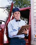 1950s 1960s MAN DRIVER DELIVERY SERVICE REPAIRMAN IN UNIFORM CAP BOW TIE HOLDING CLIPBOARD STANDING IN OPEN DOOR OF TRUCK CAB