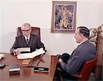 1960s MAN TALKING TO BANKER LOAN OFFICER LOANS BANK DESK