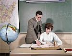 1960s COLLEGE TEEN HIGH SCHOOL COUPLE AT DESK LOOK AT NOTEBOOK GLOBE MAP CHALK BOARD BOY STANDING GIRL SEATED STUDY