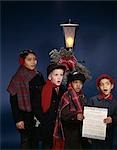 1960s GROUP MULTI-ETHNIC BOYS SINGING CHRISTMAS CAROLS UNDER LAMP POST