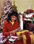 1970s SMILING YOUNG AFRICAN AMERICAN TEENAGE WOMAN WRAPPING CHRISTMAS PRESENTS TREE FIREPLACE IN BACKGROUND