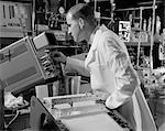 DES ANNÉES 1960 MÂLE TECHNICIEN DE LABORATOIRE VÊTU D'UNE BLOUSE DE LABORATOIRE BLANCHE LISANT UN OSCILLOSCOPE