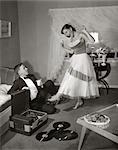 1950s 1960s TEEN COUPLE IN LIVING ROOM IN PROM DRESS & TUXEDO GUY SITTING ON FLOOR & GIRL DANCING TO MUSIC FROM A RECORD PLAYER