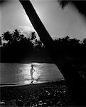 1940s SILHOUETTE OF WOMAN STANDING ON TROPICAL SHORE WITH RAY OF LIGHT COMING IN ON WATER BEHIND HER