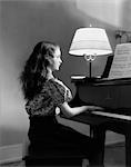 1930s SIDE VIEW OF GIRL PLAYING PIANO WITH LAMP TURNED ON NEXT TO HER