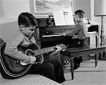 1960s PAIR OF BOYS ONE WITH BACK TO CAMERA PLAYING PIANO OTHER IN 3 4 VIEW PLAYING ACOUSTIC GUITAR