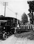 1920s DELIVERY MAN UNLOADING MILK CANS FROM LARGE TRUCK ONTO WOODEN PLATFORM ALONG SIDE OF ROAD