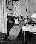 1900s TURN OF THE CENTURY BOY PLAYING AT TABLE IN SITTING ROOM WITH MOTHER IN CHAIR BEHIND HIM READING