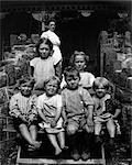 1890s 1900s TURN OF THE CENTURY GROUP OF SIX DIRTY CHILDREN SITTING ON STAIRS IN FRONT OF HOUSE WITH MOTHER STANDING ON PORCH IN BACKGROUND