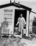 1930s MAN IN BIB OVERALLS COMING OUT OF A FARM BUILDING WITH TWO PAILS CHICKEN EGGS DOORWAY WINDOW WORKER FARMER