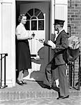 1930s POSTMAN GIVING A LETTER TO A WOMAN IN THE DOORWAY OF A COLONIAL BRICK HOME