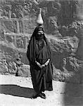 1920s 1930s EGYPTIAN WOMAN WITH PITCHER BALANCED ON HEAD WEARING TRADITIONAL ARAB MUSLIM GARB FACE VEILED