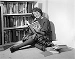 1950s 1960s SMILING YOUNG WOMAN TEEN SITTING ON FLOOR BY BOOK SHELVES READING WEARING SWEATER AND PLAID PLEATED SKIRT AND TIGHTS