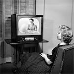 1950s TEEN SITTING WITH BACK TO VIEWER WATCHING A WOMAN LOOKING INTO A BASSINET ON HER TELEVISION IN A DARK ROOM
