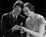 ANNÉES 1930 COUPLE SOURIANT ASSIS TÊTES PRESSÉES ENSEMBLE HOMME METTRE LA BAGUE DE FIANÇAILLES À DOIGT DE FEMME