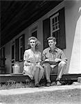 1940s COUPLE MAN WOMAN SITTING PORCH FARM HOUSE LOOKING OFF TO SIDE LEDGER BOOK MAN'S LAP SMALL MILK CONTAINER WOMAN PLAID DRESS