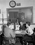 1960s DISC JOCKEY AT RADIO STATION MIKE MICROPHONE TALK WITH COUPLE MAN WOMAN AT TABLE MAN IN CONTROL ROOM