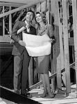 1960s COUPLE IN NEW CONSTRUCTION WITH ARCHITECTURAL PLANS