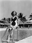 ANNÉES 1940 BRUNETTE SE BAIGNER BEAUTÉ LEANING ON PISCINE LADDER AVEC MOTEL & PALMIERS EN ARRIÈRE-PLAN