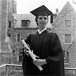 1950s SMILING FEMALE GRADUATE HOLDING DIPLOMA WITH STONE CAMPUS BUILDINGS IN BACKGROUND