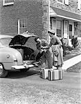 1940s SMILING COUPLE WOMAN HANDING MAN LUGGAGE TO PUT IN TRUNK OF SEDAN CAR IN DRIVEWAY BY HOME