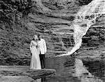 COUPLE DES ANNÉES 1970 EN TENUE HABILLÉE, DEBOUT SUR LE ROCHER À CÔTÉ DU RUISSEAU AVEC CHUTE D'EAU QUI DESCEND DERRIÈRE EUX