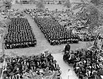 1960s VIEW FROM ABOVE STAGE AREA LOOKING DOWN ONTO 2 SECTIONS OF SEATED GRADUATES WITH SURROUNDING AUDIENCE HOLDING UMBRELLAS